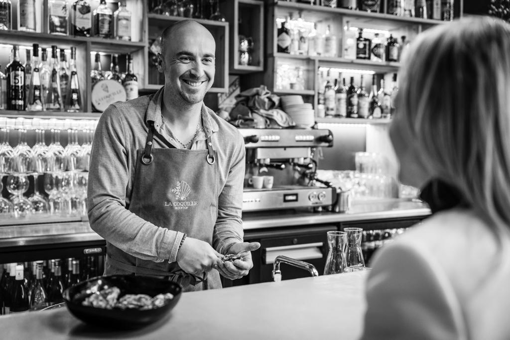 Restaurant La Coquille, Le patron du restaurant à Serre Chevalier ouvrant des huîtres fraîches pour un client satisfait dans une salle moderne